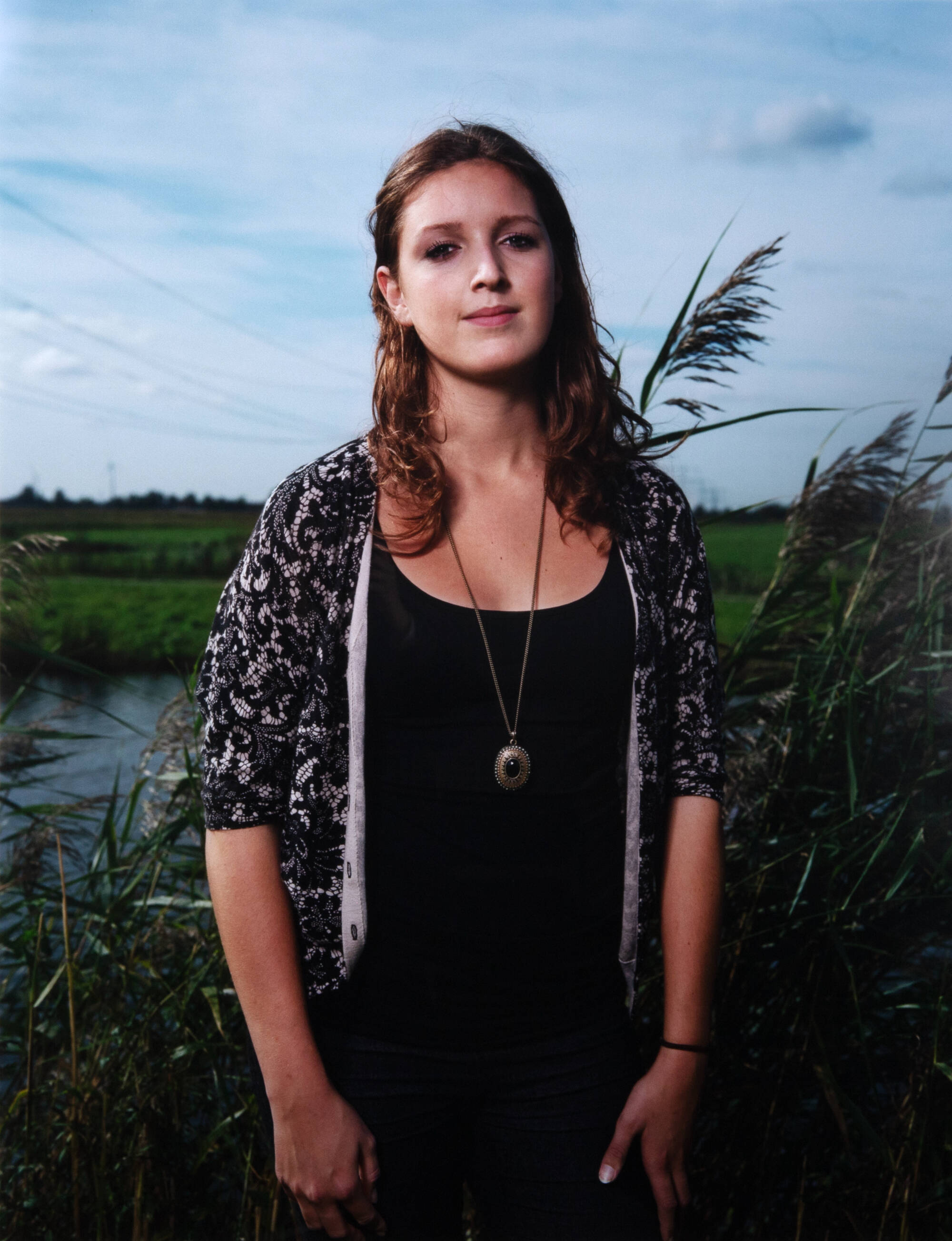 A teenager stands in front of a pond in the tall grass. They faces the camera wearing a black tank top with a black lace sweater and a long-chained gold and black necklace.